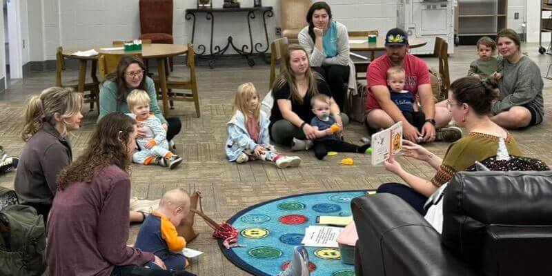 A photo of a library storytime. Moms and dads sit in the floor with their babies, looking at a librarian reading a picture book.