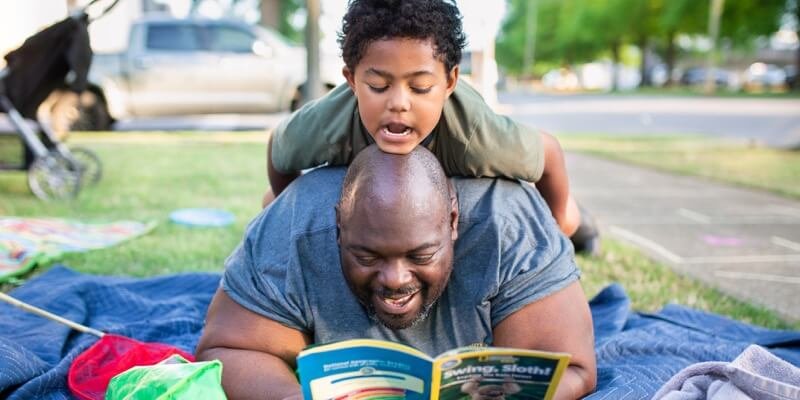 A photo of a father and son lying on a picnic blanket outdoors. The father is lying on his stomach with an open book, and his young son is lying on his back also looking at the book.