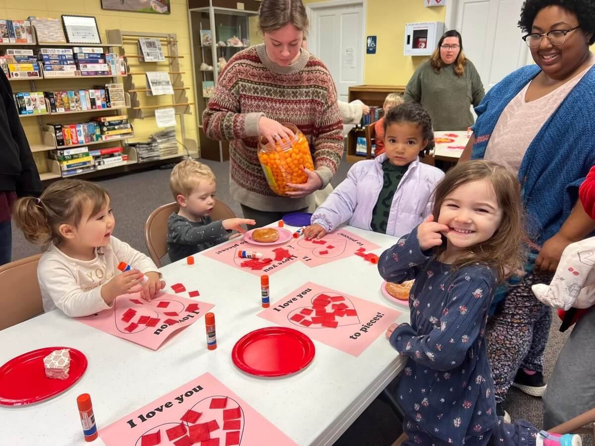 Parents and kids doing a craft activity around a table at Rogersville Public Library. One mom is helping hand out snacks.