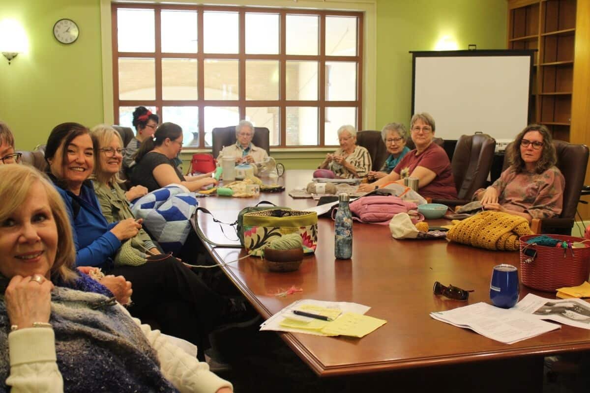 11 women of all ages sit around a table with their their knitting projects for the Close Knit Circle meeting at Fairhope Public Library