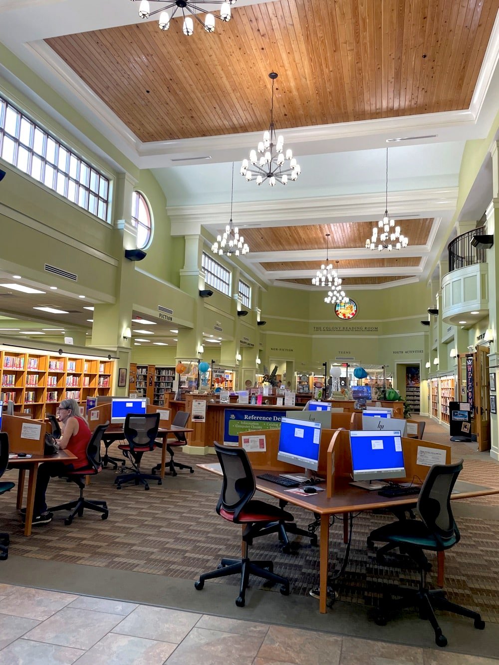 The first floor of the Fairhope Public Library. There are people using public computers, the walls are lined with book shelves, and there are chandeliers hanging from the high ceiling.
