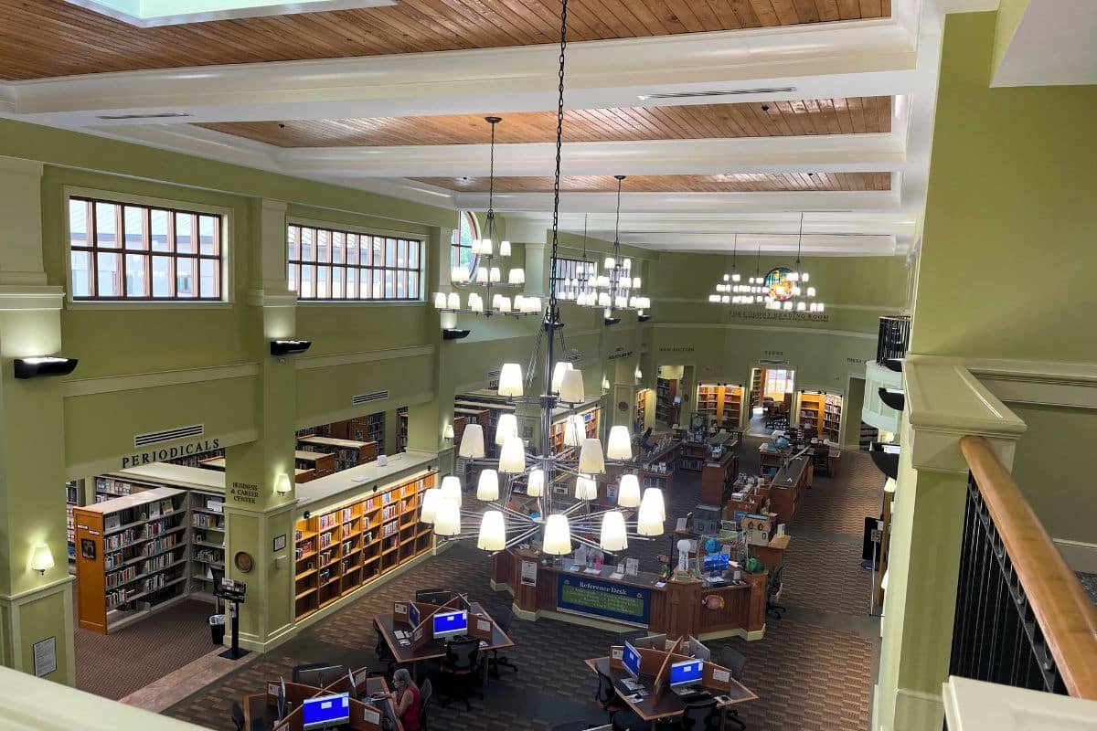 A photo overlooking the first floor of the Fairhope Public Library. Large chandeliers hang fro the tall ceilings, and you can see shelves of books, desks with public computers, and more. The ceiling is natural wood and the feel is cozy and warm.